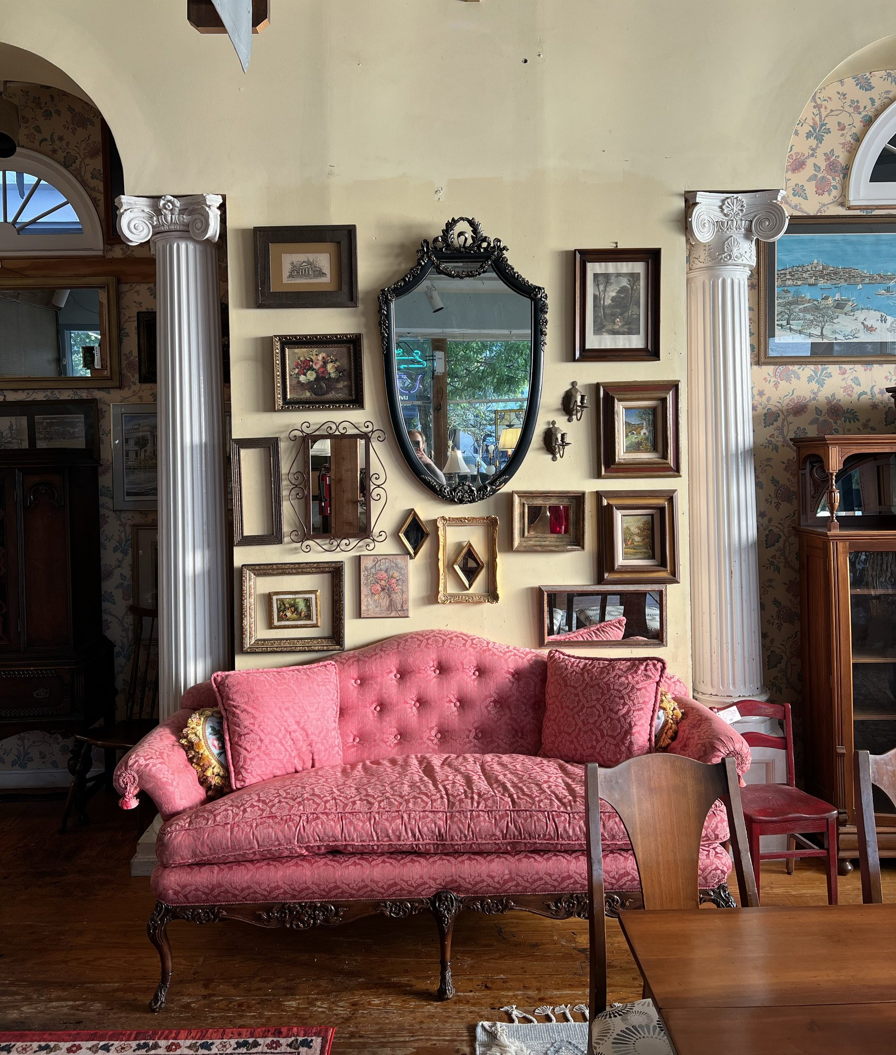 An ornate oval mirror on a gallery wall of framed art between two fluted columns, above a pink tufted sofa