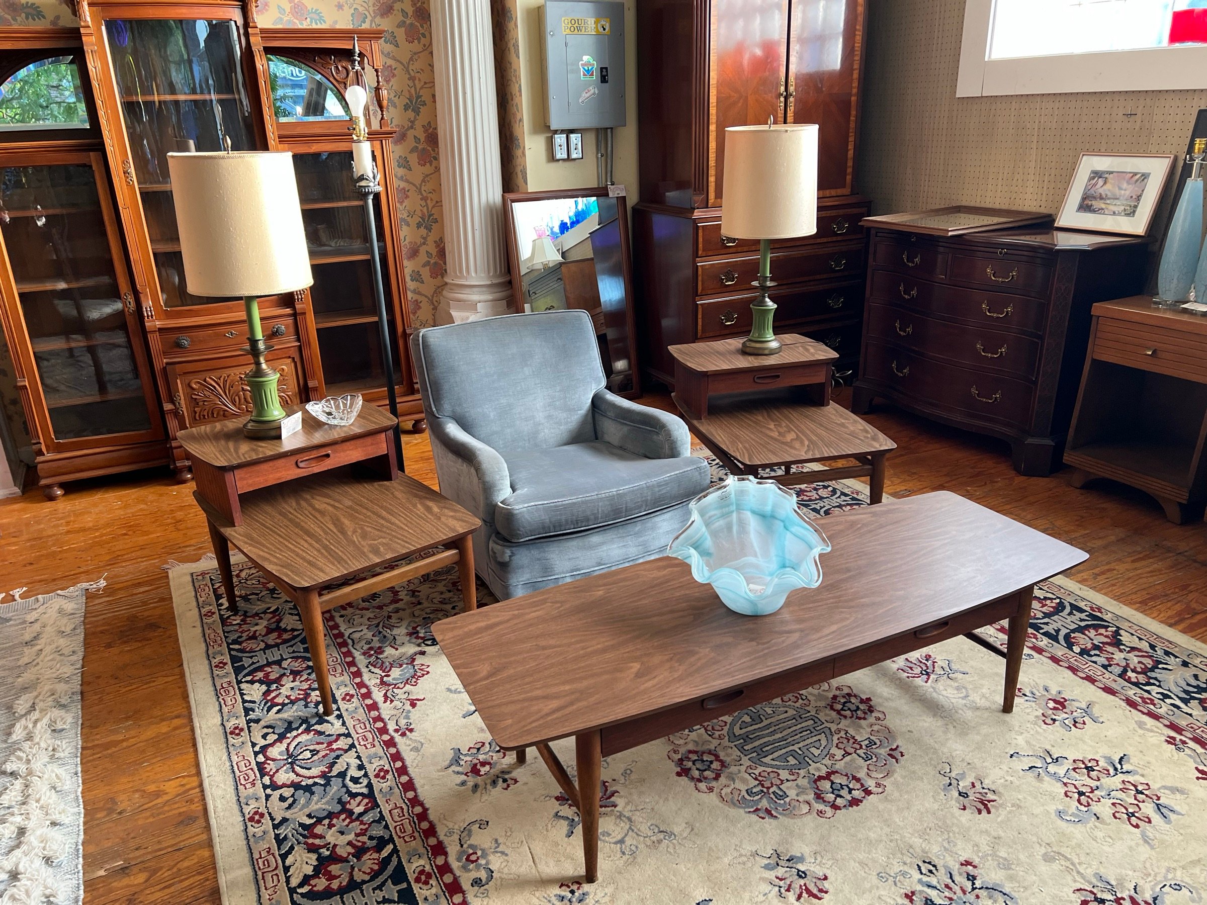 A grey lounge chair with a walnut coffee table and matching end tables, a blue art-glass bowl, and lamps on layered rugs