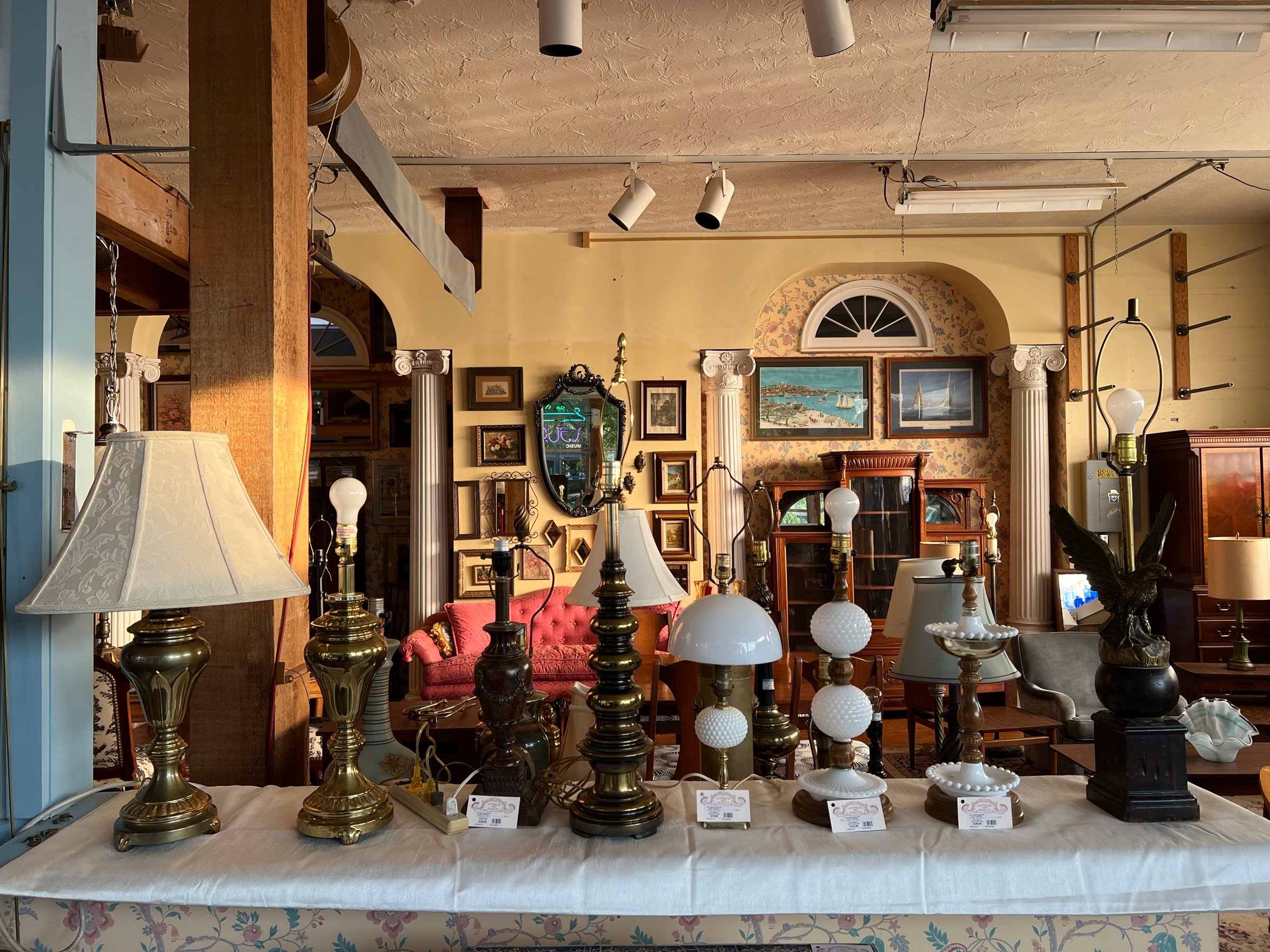 A long table lined with brass, milk-glass and hobnail table lamps, with the shop parlor behind