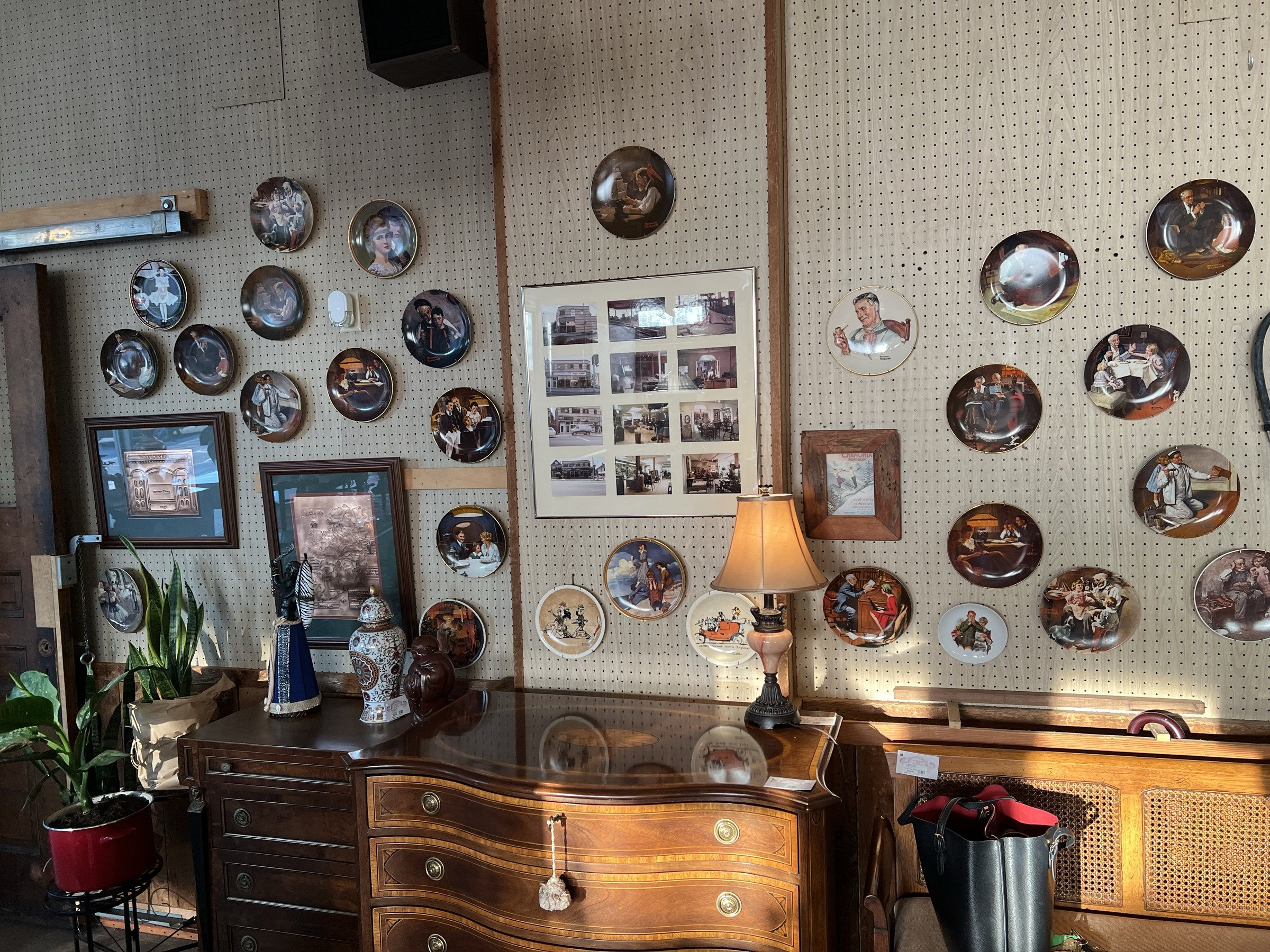 A wall of collector plates and framed photographs above a serpentine-front dresser, lit by a warm table lamp