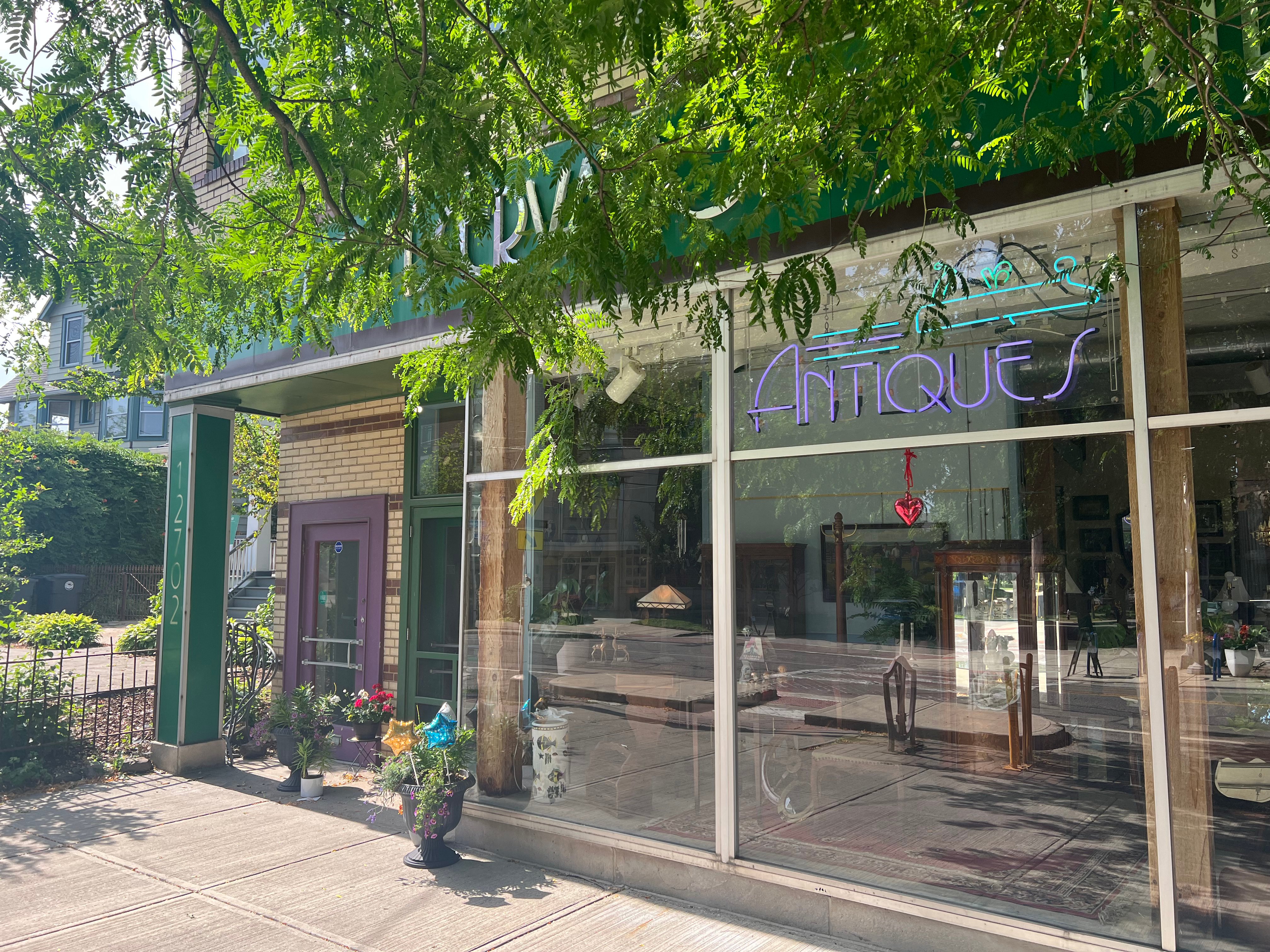 The shop's shaded storefront entrance framed by trees, with its neon Antiques sign glowing in the window and potted plants by the door